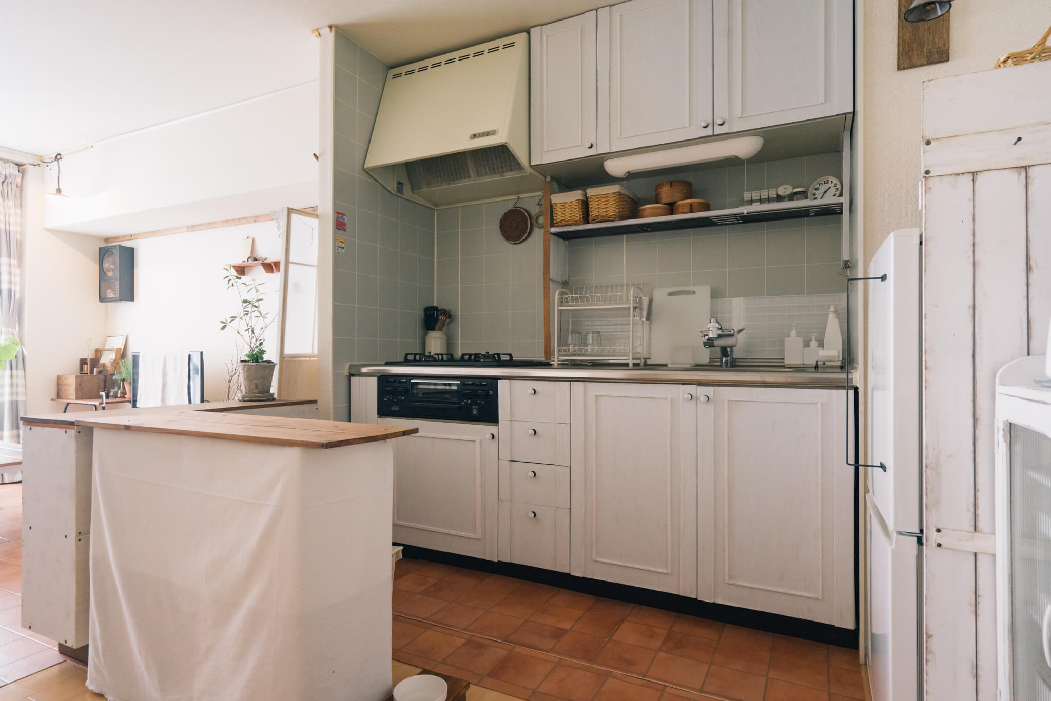 壁側に取り付けられたキッチンとリビングの間に、L字型に置かれたキッチンカウンター。普通のカラーボックスの上に天板を置き、白い布で覆っただけ。